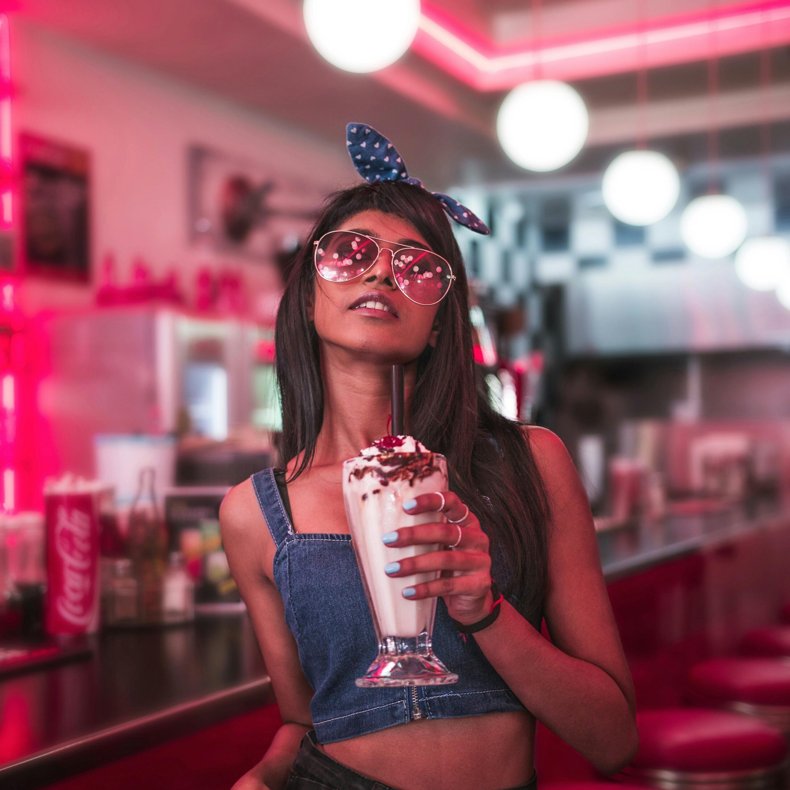 Young woman in fashionable attire enjoying a milkshake at a retro diner with vibrant lighting.